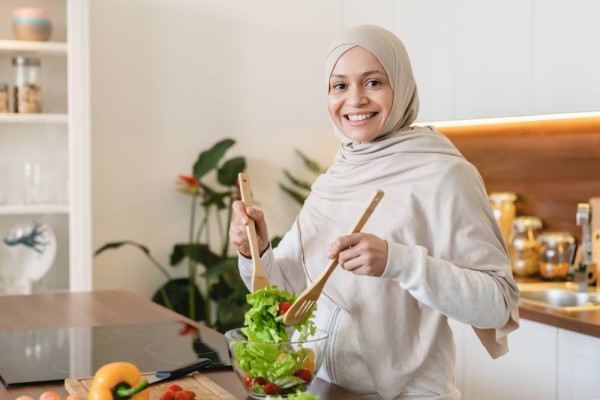 Successful mature middle-eastern wife woman in hijab cooking vegetable salad, preparing vegan vegetarian food at home in the kitchen.